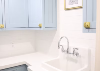 Symmetrical laundry room featuring dusty blue shaker cabinets with gold latch hardware, white quartz countertops,a vintage high back farmhouse sink.