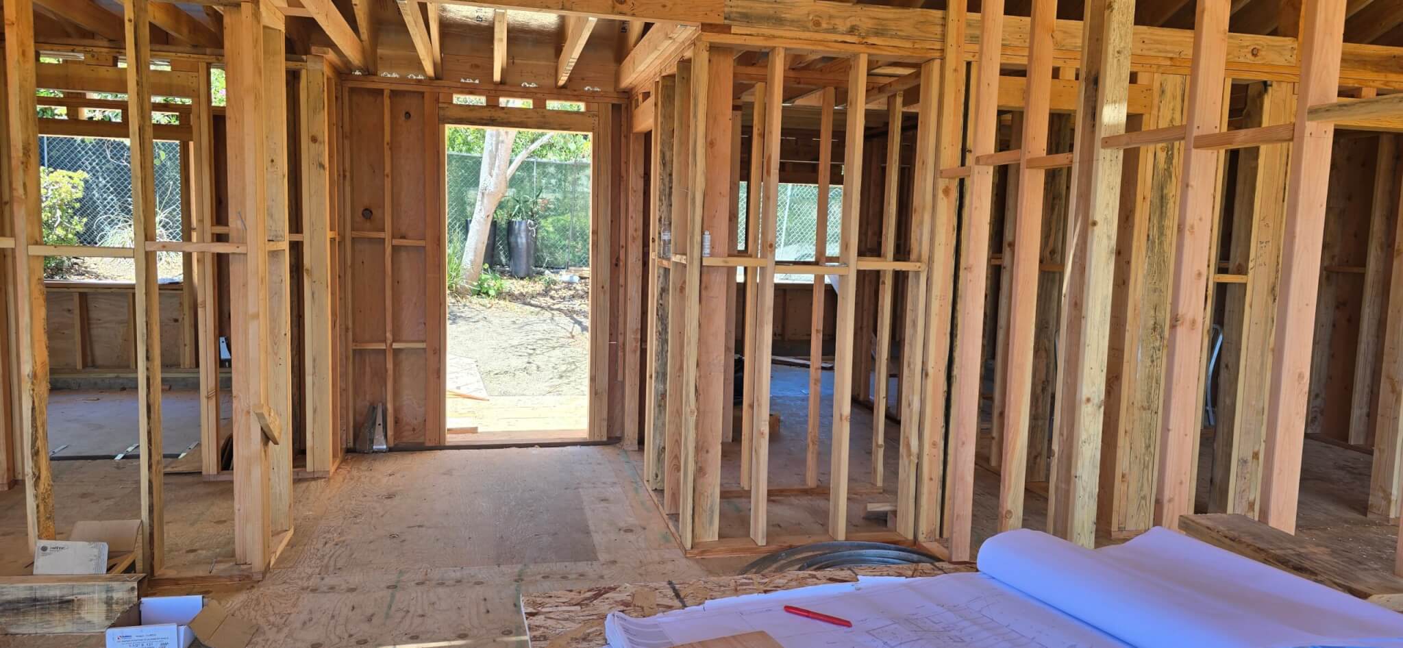 Interior view of a residential construction site in Summerland, CA, showing the raw wood wall framing, subflooring, and ceiling joists. Architectural blueprints and a red marker rest on a workbench in the foreground, looking through a framed doorway toward a sunny, tree-lined backyard.