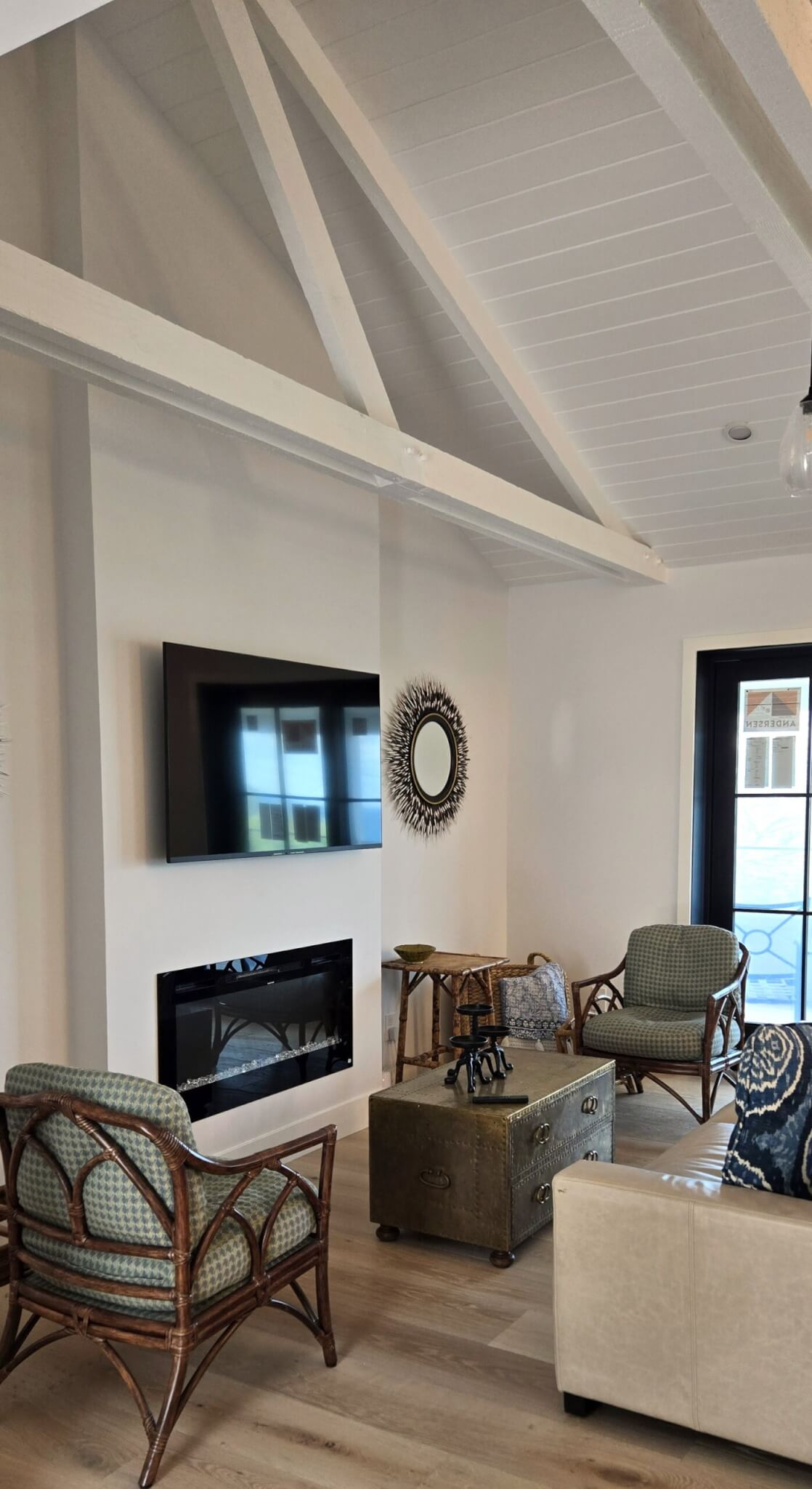 Bright coastal-style living room featuring a vaulted tongue-and-groove ceiling with exposed white beams, a wall-mounted TV over a modern linear electric fireplace, and rattan armchairs.